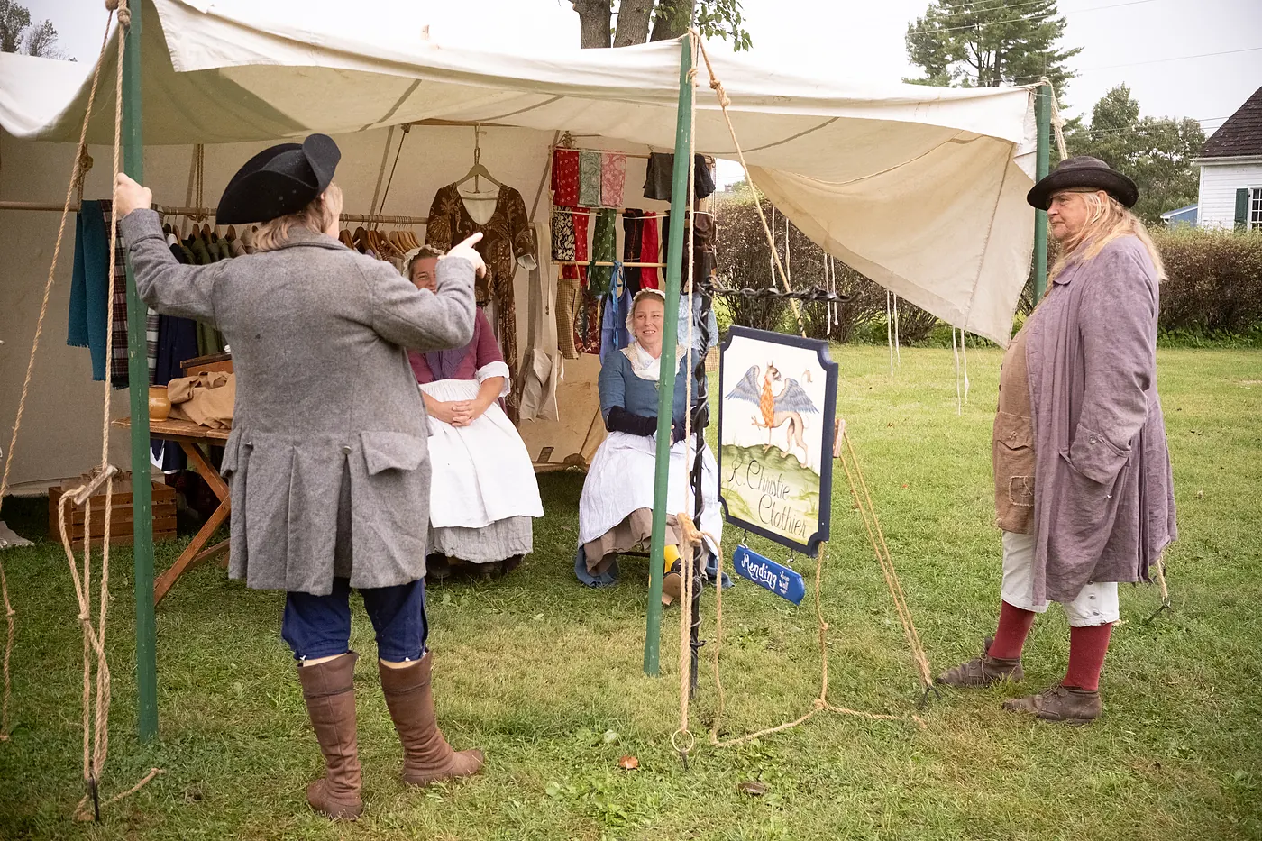 Reenactors in colonial dress talking outside a clothier's tent hung with garments