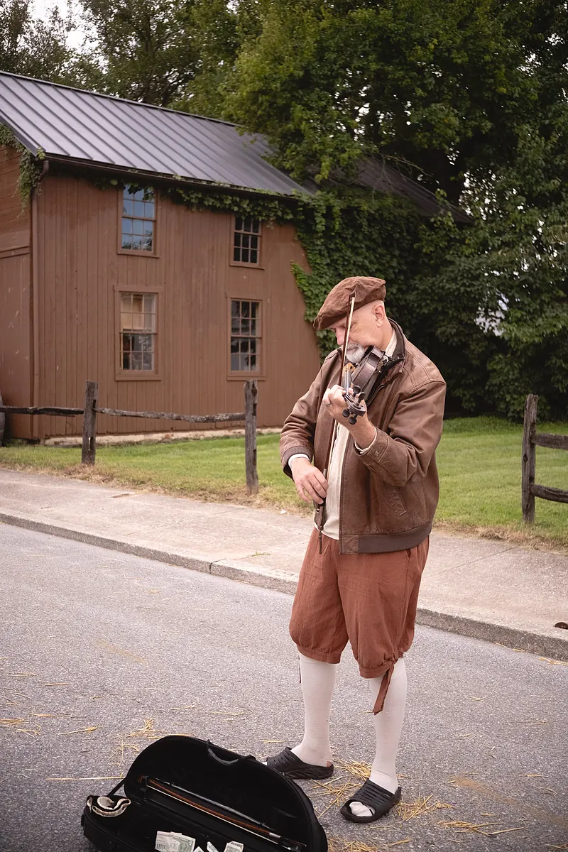 A fiddler in a flat cap and knee breeches playing in the street beside his open fiddle case
