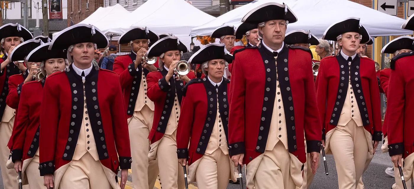 Fife and drum corps members in red coats and tricorn hats marching past the vendor tents