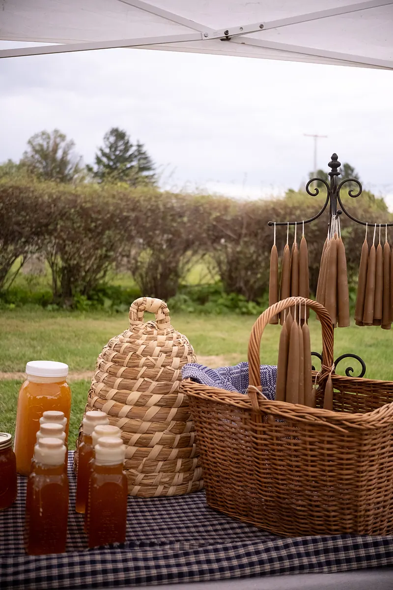 Jars of honey, a straw bee skep, and beeswax candles on a checked tablecloth