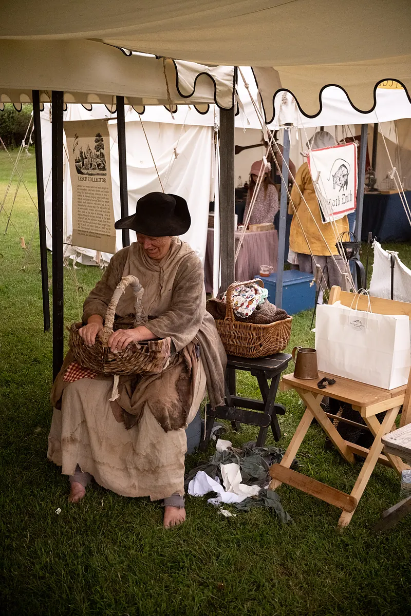 A living historian portraying a leech collector, seated with a wicker basket beneath a canvas tent